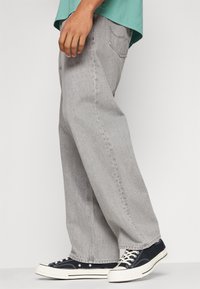 Osoba v volných světle šedých džínách, černobílých plátěných teniskách a zeleném tričku, stojící na jednobarevném světlém pozadí.