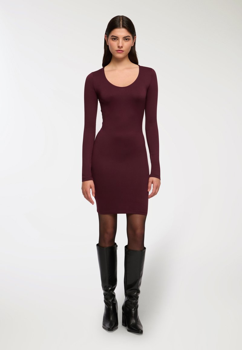 Bordowa sukienka z długim rękawem wykonana z elastycznego materiału, o dopasowanej sylwetce, zestawiona z błyszczącymi czarnymi, długimi botkami na obcasie.