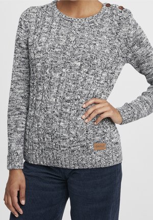 Grauer und weißer zopfgestrickter Pullover mit zwei braunen Schulterknöpfen und einem kleinen Lederpatch, getragen zu dunkelblauer Cordhose.