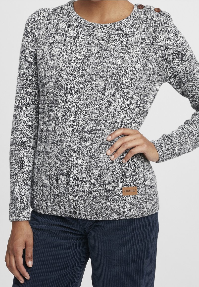 Pull à torsades gris et blanc avec deux boutons marron aux épaules et une petite pièce en cuir, porté avec un pantalon en velours côtelé bleu foncé.