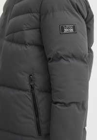 Gray puffer jacket with quilted design, featuring a side zippered pocket and a fabric tag on the upper arm with logo details.