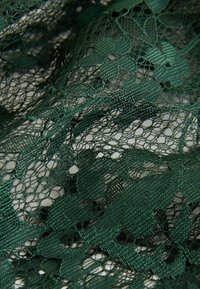 Dunkelgrüner Spitzenstoff mit floralen Mustern, filigranen offenen Geweben und einer glatten Textur, die detaillierte Stickereien und Nähte hervorhebt.