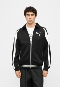 Νεαρός άνδρας φορώντας μαύρο αθλητικό μπουφάν Puma με λευκές ρίγες και λογότυπο, στεκόμενος μπροστά σε απλό ανοιχτό φόντο.