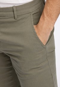 Olivgrüne Hosen aus einem glatten Stoff, mit einer Seitentasche, Gürtelschlaufen und einer taillierten Passform mit minimal sichtbaren Nähten.