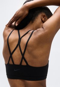 Γυναίκα που απλώνει τα χέρια πίσω από το κεφάλι, φοράει μαύρο αθλητικό σουτιέν με χιαστί λουριά στην πλάτη, σε απλό φωτεινό φόντο.