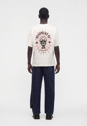 Mand set bagfra iført hvid T-shirt med kranie- og stjerne grafik i rød og sort, kombineret med vidde mørkeblå jeans og sorte sko.