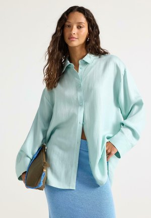 Kvinde med langt krøllet hår iført en oversized, lyseblå skjorte med knapper og en blå strikket nederdel, der holder en brun og blå taske.