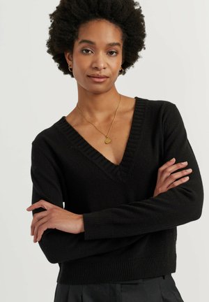 Vrouw met krullend haar die een zwarte V-hals trui draagt, gouden hoepeloorbellen en een gouden hangerketting, staand met gekruiste armen.
