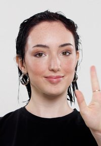 Korte mouwen zwarte shirt; nat donker haar; ringoorbellen; natuurlijke makeup; hand geheven in een zwaaigeste; lichte huid met sproeten.