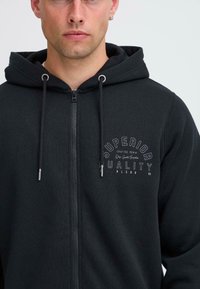 Sort zip-up hoodie lavet af blødt stof, med snoretræk i hætten og et trykt logo på brystet, samt metaldetaljer på lynlåsen.