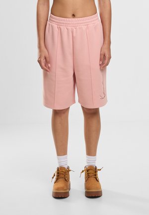 Inimene, kes kannab heleroosad põlvekõrgused lühikesed pükse, valgeid tavalisi sokke ja beeže paeltega töösaapaid, seisab ühtlase halli tausta ees.