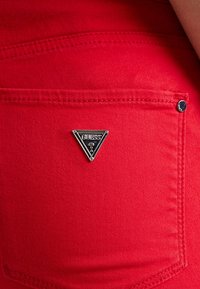 Tissu en denim rouge avec un logo triangulaire argenté "Guess" sur la poche, mettant en valeur des détails de couture et une rivet décoratif en métal.