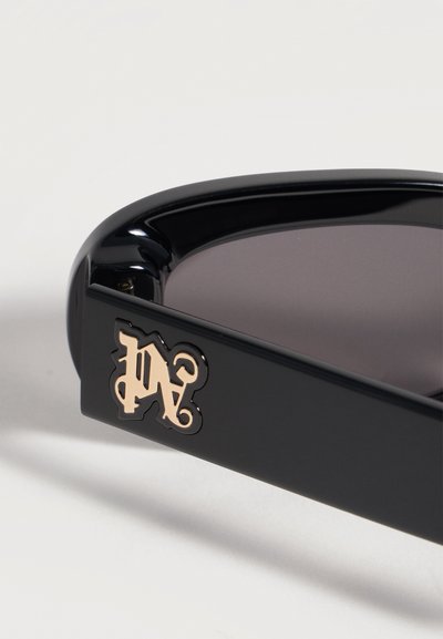 Branché de lunettes de soleil noir avec détail décoratif de monogramme doré, attaché à des verres teintés foncés sur fond blanc.