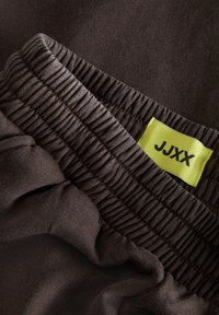 Hnědá látka s texturovaným povrchem, s elastickým pasem a světle zelenou etiketou s černým textem "JJXX." Detailní pohled.