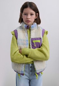 Chaleco de forro polar en crema, morado y colores pastel con cuello alto, cremallera frontal y un bolsillo con cremallera. Combinado con un suéter verde y jeans.