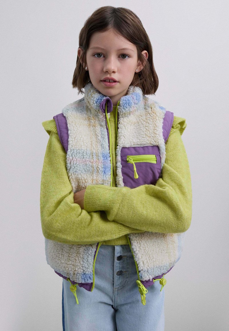Chaleco de forro polar en crema, morado y colores pastel con cuello alto, cremallera frontal y un bolsillo con cremallera. Combinado con un suéter verde y jeans.