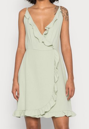 Femme portant une robe portefeuille sans manches vert clair avec un bord à volants et un tatouage floral visible sur l'épaule droite.