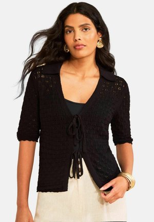 Žena s dlhými tmavými vlasmi, ktorá nosí zlaté kruhové náušnice, čierny textúrovaný pletený sveter viazaný vpredu, čierne tričko, béžovú sukňu a zlaté náramky.