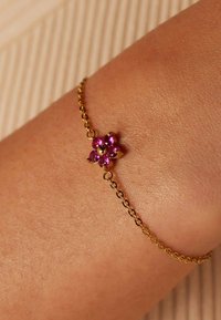 Gouden kettingarmband met een bloemmotief en vijf roze edelstenen in het midden, geplaatst tegen een gestructureerde huidachtergrond.