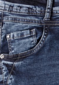 Jean en denim avec un délavage bleu foncé, des coutures en contraste et une petite poche fixée par un bouton en argent près de la taille.