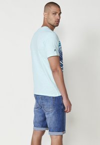 Ljusblå kortärmad T-shirt med ett mönstrat tryck på vänster sida; som sätts ihop med denimshorts med uppvikta ärmar. Mjuk, texturerad tyg.