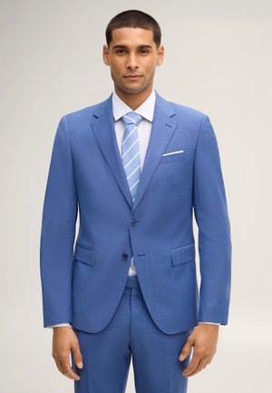 Man draagt een lichtblauw pak met een wit overhemd en een blauw gestreepte stropdas, staand tegen een effen lichte achtergrond.