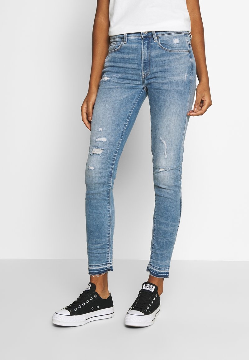 Jeans ajustados de mezclilla azul claro, con elementos desgastados; combinados con zapatillas de lona negras con suelas blancas y cordones negros.