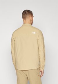 Beżowa koszula zapinana na guziki z długimi rękawami, miękką teksturą i logo na plecach. Posiada kołnierz typu spread oraz regulowane mankiety.