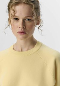 Sweatshirt jaune clair avec un col rond côtelé, des manches raglan et une texture douce, offrant une coupe décontractée.