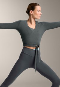 Top grigio a coste con maniche lunghe, indossato sopra un reggiseno sportivo grigio, abbinato a leggings grigi a vita alta con dettaglio a fiocco in vita.
