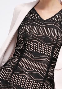 Abito nero a fantasia con ritagli geometrici, caratterizzato da una scollatura a V e un design aderente. Indossato sotto un blazer rosa chiaro.