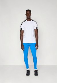 White athletic t-shirt with black striped accents on shoulders, paired with blue slim-fit pants featuring white stripes on the sides. Black shoes.