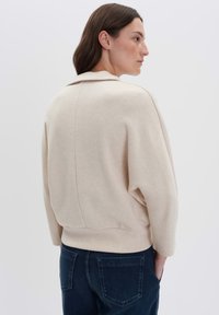 Frau mit langen dunklen Haaren, die einen beigen Pullover und dunkle blaue Jeans trägt, steht leicht zur Seite gedreht vor einem einfarbigen weißen Hintergrund.
