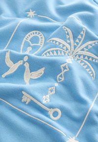 Tessuto azzurro chiaro con ricami bianchi raffiguranti uccelli, una palma, forme geometriche e un motivo a chiave, caratterizzato da una notevole texture e dettagli.