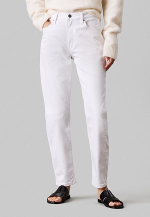 Person wearing white straight-leg jeans, beige fuzzy sweater, and black open-toe flat sandals, standing against gray background.