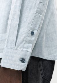 Hellblaues Hemd mit Fischgrätenmuster, langen Ärmeln, blauen Knöpfen an den Manschetten und strukturiertem Stoff.