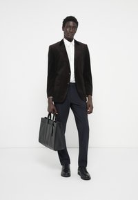 Schwarzer Cord-Blazer, weißes Hemd, marineblaue Hose und schwarze Lederschuhe; hält eine strukturierte schwarze Ledertasche.