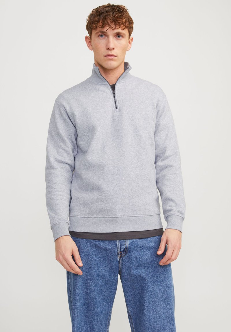 Sweat-shirt gris à col moitié zip, fabriqué en tissu doux. Il présente des poignets et un ourlet côtelés, porté par-dessus un t-shirt noir et un jean bleu.