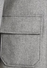 Tissu gris en mélange de laine avec un motif chevrons. Comprend une poche plaquée rectangulaire aux bords arrondis et une couture apparente.