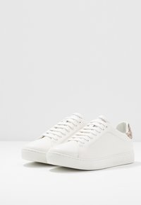 Ett par rena vita låga sneakers med vita snören och beigefärgade mönstrade hälar på en vit yta.