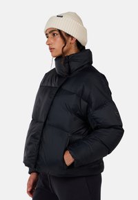 Giacca a vento nera con colletto alto, tasche con zip e design trapuntato. Indossata con un berretto di maglia crema. Texture liscia e lucida.