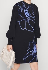 Abito felpa nero con maniche oversized e un grande motivo floreale blu. Il modello tiene una borsa a tracolla di colore crema con texture.