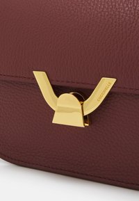 Burgundy leather handbag featuring a glossy gold-tone clasp with a unique geometric design and embossed brand name detail.