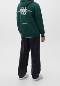Sudadera verde con estampado gráfico blanco en la parte posterior, combinada con pantalones negros holgados y zapatillas blancas. Tejido suave y texturizado.