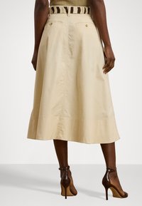 Beige mid-length skjørt med en utvidet design, sidelommer og subtile sømdetaljer, kombinert med brune sandaletter med høye hæler.