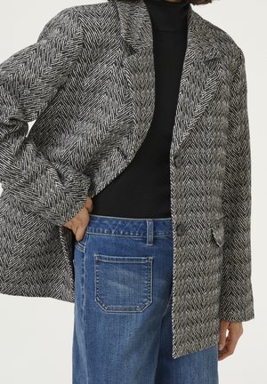 Femme portant un col roulé noir, un blazer gris à motifs avec boutons, un jean taille haute bleu avec poche arrière, main dans la poche, fond neutre.