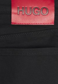 Tela negra con una textura suave que presenta una etiqueta rectangular roja grabada con la palabra "HUGO" en negro.