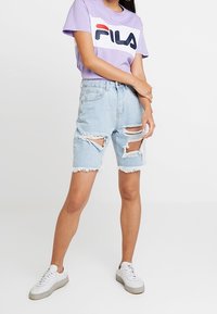 Light blue ripped denim shorts with frayed hem, paired with a lavender t-shirt featuring a bold, colorful FILA logo, and white sneakers.