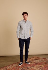 Een knoopshirt met een multicolore bloemenpatroon, lange mouwen en een kraag. Gekleed met marineblauwe broek en blauwe sneakers op een patterned tapijt.