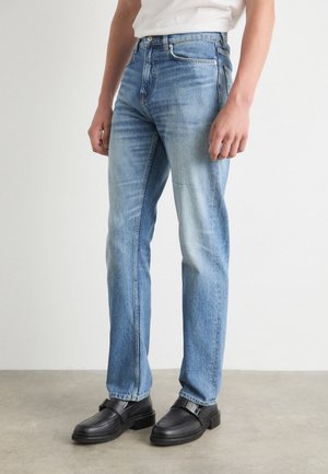 Muž v modrých džínách s rovnými nohavicemi, bílém tričku a černých kožených mokasínách stojí na šedé podlaze před jednobarevnou bílou zdí.